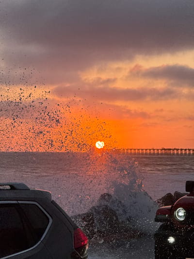 The image shows a dramatic sunset over the ocean in Oceanside, California. The sun, a bright orb of orange, is partially obscured by a wave crashing in the foreground, creating a spectacular spray of water. The sky is filled with vibrant hues of orange, pink, and purple, with scattered clouds. A pier stretches across the horizon, lined with streetlights and silhouetted figures. In the foreground, two vehicles, a grey SUV and a red Jeep, are parked near the water, partially engulfed in the spray from the waves. The scene captures the raw power and beauty of the ocean at sunset.