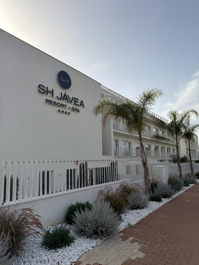 The image depicts the exterior of the SH Jávea Resort & Spa, a modern, white building with multiple stories and balconies. The resort is located in Les Duanes De La Mar, Spain, as indicated by the signage on the building. Palm trees are visible in front of the building, and the surrounding area is landscaped with white pebbles and various types of shrubs and grasses. A brick-paved walkway runs alongside the property. The sky is overcast with some clouds. There are no people visible in the image. The overall scene suggests a clean, contemporary resort environment.