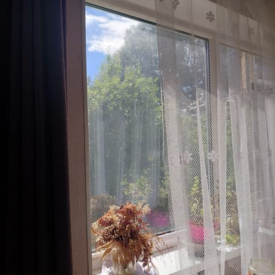 The image depicts a close-up view of a window in Skuķīši, Latvia. On the left, a dark brown curtain is drawn partially closed. To the right of the curtain, a sheer white lace curtain with a floral pattern is visible, covering the window. Through the lace curtain and the window, lush green trees and a bright blue sky with white clouds are visible, suggesting an outdoor scene. On the windowsill, in front of the window, sits a glass vase containing a dried bouquet of flowers and grasses. The light filtering through the window illuminates the scene, creating a soft and slightly hazy effect on the glass, possibly due to dust or water droplets. The overall impression is one of domestic tranquility with a view of nature.