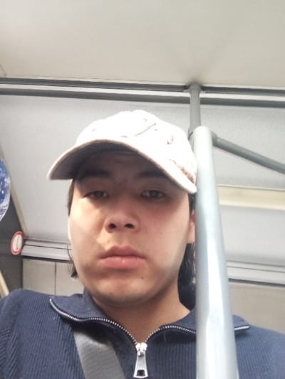An individual, identified as a young adult male, is depicted in a close-up, frontal self-portrait within what appears to be the interior of a public transport vehicle. The subject wears a light-colored baseball cap, a dark blue ribbed zip-up top with a silver zipper, and a gray seatbelt strap is visible across his chest. Small white earbuds are visible in both ears. His expression is neutral. The background shows a light-colored ceiling, a vertical gray metal grab pole on the right side, and a horizontal gray bar above it. A reflective surface and a partial red and white circular object are discernible on the far left. The specific city location of Urtaaul, Uzbekistan, cannot be confirmed from the visual information present in the image.