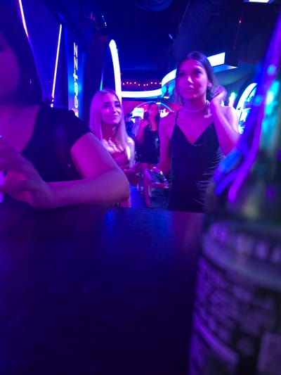 The image depicts a dimly lit, vibrant nightclub scene, likely in Leipzig, Germany, illuminated by an abundance of neon blue and purple lights. In the foreground, a person is seated at a dark, reflective surface, their arms resting on it. To their right, a blonde woman stands, smiling and looking towards the right of the frame. Behind her, another woman in a black dress is partially visible, her attention also directed away from the camera. The background reveals more of the club's atmosphere with glowing vertical light structures and other patrons. The overall impression is one of a lively evening event, with the prominent use of colored lighting creating a dynamic and immersive environment.