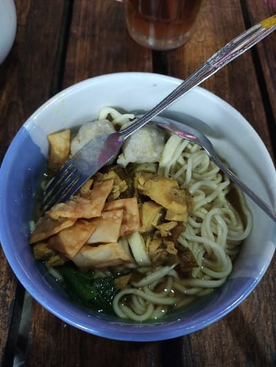 An overhead shot captures a bowl of Indonesian noodles, likely Mie Ayam or Mie Bakso, placed on a dark wooden table in Kecamatan Pandaan, Indonesia. The round bowl features a white interior and a light blue rim. Inside, a generous serving of thin, pale noodles rests in a light brown broth.
Accompanying the noodles are various toppings: chunks of what appears to be shredded or diced chicken, several pale, round meatballs (bakso), pieces of green leafy vegetables (possibly bok choy or mustard greens), and golden-brown, crispy fried wonton skins. A metal fork and spoon are partially submerged in the bowl, suggesting the meal is either about to be eaten or is in progress.
To the left of the bowl, a portion of a glass containing a amber-colored beverage, possibly iced tea, is visible. The wooden table has a rustic appearance with visible grain and gaps between the planks. The lighting is dim, indicating an indoor setting or an evening/nighttime meal. No text is visible in the image.