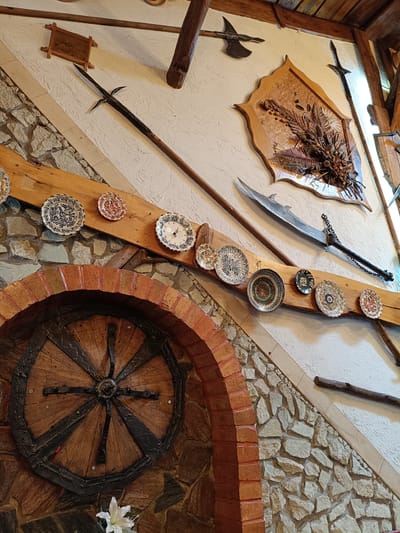 The image depicts a wall decorated with various items in what appears to be a rustic or medieval-themed interior. Dominating the scene are several weapons, including two spears, a sword with a chain attached, and an axe. There are also numerous decorative plates, some with intricate blue and white patterns, and others with red and white designs. A wooden wheel, likely part of a cart or wagon, is visible in the lower left corner, framed by a brick arch. A bouquet of dried flowers and grasses is mounted on the wall. The wall itself is a mix of textured white plaster and rough-hewn stone. This scene is located in Câmpia Turzii, Romania.