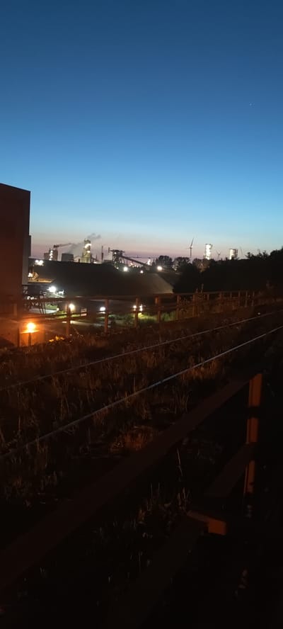 The image depicts an industrial landscape at dusk in Salzgitter, Germany. In the foreground, there are railway tracks partially obscured by dry grass and foliage. A wooden fence runs along the tracks. To the left, a portion of a large, dark structure, possibly a building or a storage facility, is visible. Further in the midground, a large mound of dark material, likely coal or slag, is prominent, with several industrial structures and a crane visible on top. In the distance, the lights of a factory complex illuminate the scene, with plumes of steam or smoke rising from some of the buildings. Several wind turbines are also visible against the twilight sky. The sky transitions from a deep blue at the top to lighter shades of blue and pink near the horizon. There are no people visible in the image. The overall impression is one of an active industrial area at the end of the day.