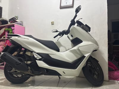 This media file is a full-shot, eye-level photograph of a white Honda PCX 160 scooter parked indoors, likely in a residential setting in Cibinong, Indonesia. The scooter is the primary subject, positioned in the center of the frame, angled slightly towards the viewer's left. It's a bright white scooter with black accents, including the seat, handlebar grips, mirrors, and the exhaust system. The scooter's "PCX 160" logo is visible on the rear bodywork.
The background consists of a plain white wall, typical of an indoor space. To the left of the scooter, a pink shelving unit is partially visible, filled with various items including a black helmet, stuffed toys (a green alligator and pink plush toys), and a patterned bowl. A framed certificate or document hangs on the wall above the shelving unit. To the right of the scooter, a dimly lit doorway or alcove is visible, with some furniture and stacked items, possibly luggage or bedding, covered in pink and black patterned fabric. The floor is tiled with a light-colored pattern.
There is no apparent activity or event taking place; the scooter is simply parked. The lighting suggests it is daytime, though the indoor setting and some shadowed areas create a slightly dim ambiance. The overall impression is that of a scooter parked in a personal storage space or garage. No specific emotions are conveyed. The only visible text is the "PCX 160" logo on the scooter.