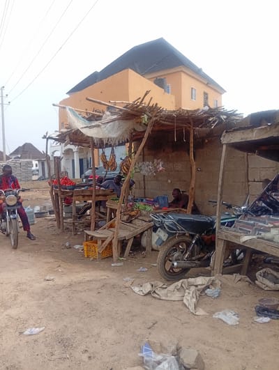 The scene depicts a roadside market in what appears to be Aviation Quaters, Nigeria. The market consists of makeshift stalls constructed from wood, bamboo, and corrugated metal. One stall displays tomatoes and plantains, while another seems to offer various goods, including eggs and snacks. People are present at the stalls, presumably vendors. Two men are at one stall. A man on a motorcycle is on the road. Several motorcycles are parked near the stalls. The ground is sandy and littered with debris. In the background, there are buildings, including a two-story structure with an orange facade and a dark roof. Power lines are visible in the sky.