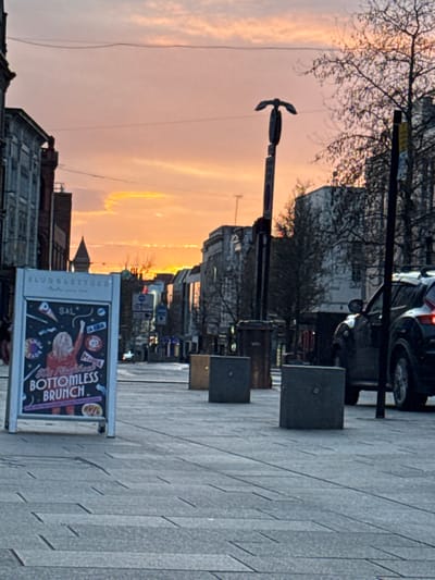 The image depicts an urban street scene in Preston, United Kingdom, at dusk or dawn, characterized by a vibrant orange and pink sky. The perspective is from a paved pedestrianized area, likely Lancaster Road, looking eastward.
In the immediate foreground on the left, a white A-frame pavement sign stands upright. The sign's graphic advertises "Mamma Mia's Bottomless Brunch" for "SLUDGE & LETTUCE," featuring an illustration of a blonde-haired individual raising a drink amidst various icons. The paved ground consists of grey rectangular blocks.
Moving into the midground, a line of three square, grey concrete bollards with metallic centers is positioned on the paved surface, extending towards the right. To their right, a dark-colored SUV, possibly a Volvo, is partially visible, parked or stopped with its rear right quarter and illuminated taillight facing the viewer. Adjacent to the bollards and further back, a tall, dark street pole with a distinctive, multi-pronged fixture on top stands, with a dark brown rubbish bin at its base. Another similar bin is visible further right. Bare trees line the right side of the street, contrasting with multi-story buildings that display older architectural styles.
In the background, the street extends, flanked by more buildings. A building with a conical or spire-like roof is discernible on the left side, further down the street. Overhead, thin utility wires stretch across the sky. No human figures are present or clearly discernible in the scene.