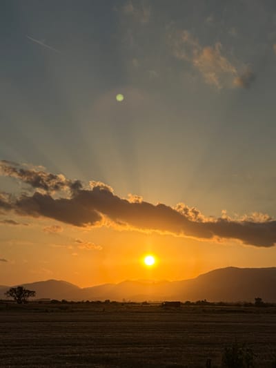 A sunset over a rural landscape in Blatets, Bulgaria. The sun is a bright orange disc low on the horizon, casting golden rays through dramatic clouds. In the foreground, a harvested field stretches towards a line of silhouetted mountains. A solitary tree stands to the left of the frame, and a small building and distant trees are visible on the horizon. The sky transitions from a soft blue at the top to a warm orange near the horizon, with a faint green circular anomaly visible in the upper sky, possibly a lens flare or atmospheric phenomenon. A thin white contrail from an aircraft is visible in the upper left quadrant of the sky.