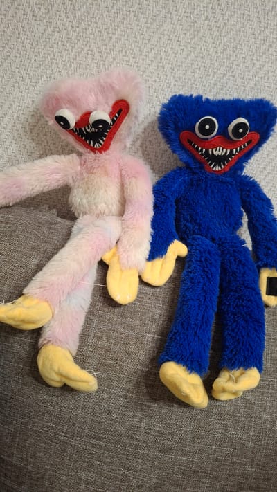 Two plush toys, a blue one and a pink one, lie on a textured surface. The blue toy is positioned above the pink one and is mostly in focus. Both toys are a fuzzy, furry material and resemble cartoon characters with large, round eyes, red mouths filled with sharp, black and white teeth, and yellow hands and feet. The blue toy has a black rectangular detail on its upper body. The pink toy has a gradient of pink and white fur. The background surface appears to be a sofa or upholstered furniture, with a lighter, textured fabric underneath the toys on the left side of the image. The lighting suggests an indoor setting, possibly during the day, with no specific time of day or weather conditions being discernible. No text is visible in the image. The scene is static, with the toys simply posed.