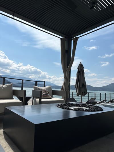 An outdoor patio with covered seating overlooks a scenic vista of water and mountains under a blue sky filled with fluffy white clouds. The patio features modern furniture, including chairs with cushions, a closed parasol, and a fire pit. The architecture has a contemporary design with sleek lines and dark framing. The scene evokes a sense of luxury and relaxation.