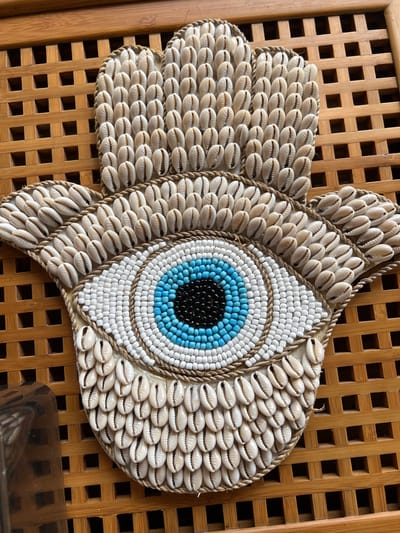 A close-up shot depicts a decorative fish crafted from seashells and beads, placed against a backdrop of a light brown wooden lattice structure. The fish's body is predominantly made of small, white cowrie shells. The eye of the fish is composed of concentric circles of black, blue, and white beads. A braided twine outlines the fish and its various sections. A small section of a shiny, dark object is visible in the lower right corner. The location is identified as Singakerta, Indonesia, based on the query.