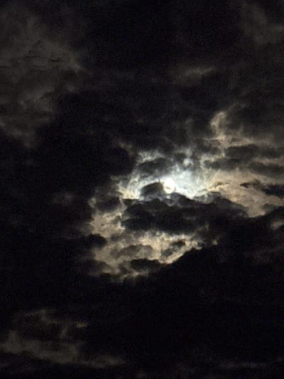 This image captures a nocturnal scene of the moon partially obscured by clouds. The main subject is the moon, visible as a bright, circular disc in the upper-middle portion of the frame, emitting a soft, diffused light. It is surrounded and partially covered by dense, textured clouds.

The clouds are varied in their appearance; some areas are dark and heavy, suggesting thick coverage, while others are illuminated by the moonlight, appearing as swirling masses of light grey and white. This interplay of light and shadow creates a dramatic and ethereal atmosphere in the night sky. The scene is set outdoors, depicting a sky at night with significant cloud cover.

Notable details include the time of day, which is clearly night, and the weather conditions, indicating a cloudy sky. The illumination from the moon suggests it might be a full or near-full moon, strong enough to shine through the breaks in the cloud layer. The overall image has a somewhat grainy texture, which is typical for low-light photography and contributes to the mysterious quality of the scene. No specific objects or activities beyond the celestial observation are depicted. There is no visible text in the image. This sky was observed in Strazhitsa, Bulgaria.