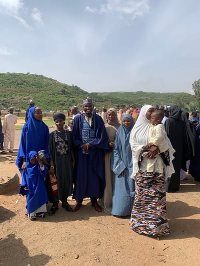 A horizontal, eye-level shot shows a group of about 10 individuals standing in a line on a dirt surface in Naraguta Hausawa, Nigeria. The landscape includes a hill covered with green vegetation and a cloudy sky. The people in the line are dressed in various traditional and religious attires, primarily in colors like blue, white, black, and patterned fabrics. Some are wearing head coverings. At least one person is holding a child. The lighting is bright and sunny, casting shadows on the ground. The overall setting appears to be outdoors, possibly on the edge of the village or town.