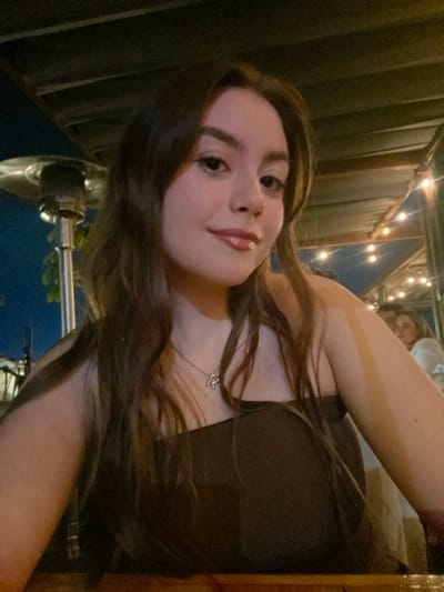 A young woman with long brown hair is taking a selfie. She is wearing a dark-colored top and a necklace with a heart pendant. She has fair skin and is looking at the camera with a slight smile. In the background, there is a person sitting and string lights. The setting appears to be an outdoor dining area in the evening, featuring metal beams. The exact location within Oaxaca City, Mexico, cannot be determined from the image alone.