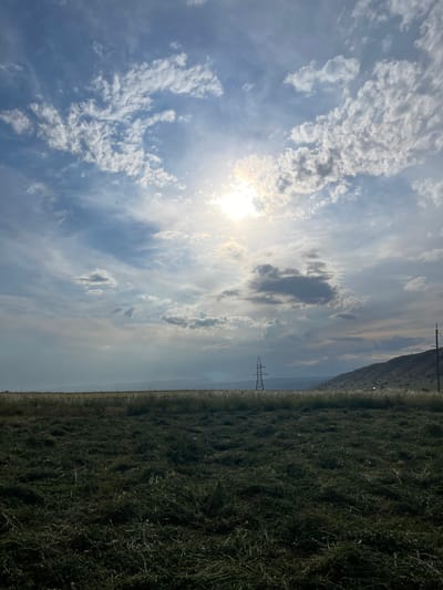 A vast, green field, recently mowed, fills the foreground. In the middle ground, a power line tower stands prominently. Beyond that, rolling hills recede into the distance. The sky above is a canvas of blue, adorned with scattered white clouds, with the sun breaking through, casting a bright glow. The scene appears to be located in Taran-Bazar, Kyrgyzstan.