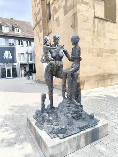 A bronze sculpture depicting a woman holding a child and a second child standing beside her is situated on a cobblestone plaza in front of a building with a stone facade in Heilbronn, Germany. The sculpture is located in the city center, likely in the vicinity of the marketplace or a pedestrian zone.

The sculpture itself is abstract and textured, with the figures appearing somewhat weathered. The woman is centrally positioned, cradling a smaller child in her arms. To her right stands a slightly taller child, leaning against the woman and looking forward. The figures are all rendered in a rough, organic style. The sculpture rests on a concrete base, which is itself placed on the paved ground.

In the background, to the left of the sculpture, is a modern building with a gray facade and a large black sign that reads "WE SUIT YOU!" with the word "PURSUITS" below it. This building appears to be a retail establishment, possibly a clothing store, with mannequins visible in the shop windows. Further down the street, a bicycle is parked. To the right of the sculpture, the stone facade of a larger, older building rises, characterized by its light brown and pinkish stone blocks. The sky above is bright and partly cloudy, suggesting daytime. The overall impression is of a public art installation in an urban setting.