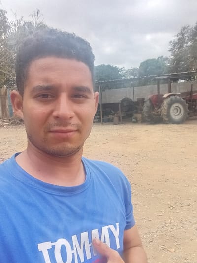 The image captures an adult male taking a selfie outdoors in a rural or industrial yard within Cujisal, Venezuela, under an overcast sky. The man, centered in the foreground, has short dark curly hair and light facial stubble. He wears a blue crew-neck t-shirt with the word "TOMMY" emblazoned in white capital letters across the chest. His right hand is visible below his chest, with the thumb extended upwards and the index finger slightly bent in a pointing gesture. In the mid-ground, behind the subject, a basic open-sided structure with a corrugated metal roof houses various pieces of machinery and equipment. Adjacent to this structure, a red tractor with prominent rear wheels is parked on the light brown, stony dirt ground. A dense line of green trees and foliage forms the background horizon.