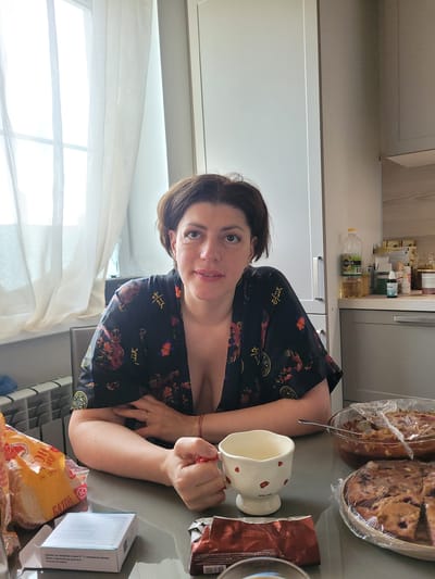 A medium-shot indoor photograph captures a woman with short, dark hair sitting at a table in what appears to be a kitchen or dining area. She is wearing a dark robe with a floral and dragon pattern, which is open at the neck, revealing cleavage. Her skin tone is fair. The woman is looking directly at the camera with a neutral expression.
In front of her on the table are several items: a white mug with a red heart pattern, a package of bread, a small white box, a red-wrapped item, and two baked goods covered in plastic wrap. One of the baked goods is a pie with fruit filling, and the other appears to be a cake or tart, also with fruit. A spoon is visible in the pie.
To the left of the woman, a radiator is partially visible. Behind her, a large window covered with sheer white curtains is present. The background also includes kitchen cabinets and shelves stocked with various items, including bottles and jars. The overall lighting suggests it is daytime. The image does not contain any elements that would place it specifically in the city of Myslyatino, Russia; it depicts a generic domestic interior.