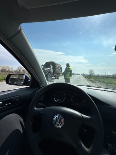 The image depicts a first-person view from the driver's seat of a left-hand drive Volkswagen car, stopped or slowing on an asphalt road in Manastir, Bulgaria. Ahead, a uniformed individual stands in the center of the two-lane road, facing away from the camera. This person wears military fatigues, a cap, and a high-visibility reflective yellow-green vest, with a long weapon slung across their back. Directly beyond this individual, a dark green military-style truck, heavily camouflaged with netting, is positioned on the right side of the road. A partial reflection of the truck's rear and camouflage is visible in the car's left exterior side mirror. The road itself has a white forward arrow painted on its surface. The environment features a clear blue sky with sparse clouds, green grassy verges, and a distant line of trees with white blossoms on the left. The car's dashboard, steering wheel with a VW logo, and the driver's left leg in dark trousers are visible in the foreground, with the digital dashboard display showing "1036."