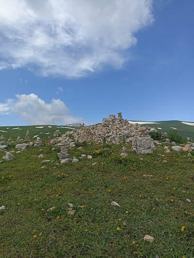 The image depicts a natural landscape, likely in the city of Podgorny, Russia, characterized by a grassy meadow dotted with small yellow flowers. In the foreground, numerous stacks of stones, or cairns, of varying sizes are arranged on the grass. Some cairns are tall and slender, while others are more spread out and low to the ground. A large pile of stones forms a prominent mound in the mid-ground, with smaller stone arrangements surrounding it. In the background, rolling green hills ascend, with patches of snow visible on their upper slopes, suggesting a higher elevation or cooler climate. The sky above is a vibrant blue, adorned with scattered white clouds, some of which are wispy and others more substantial. The overall scene evokes a sense of remoteness and natural beauty.