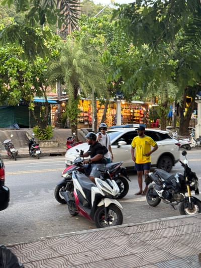 The image depicts a street scene in Ban Klong Haeng, Thailand. Several motorcycles are parked along the side of a road, with some people standing near them. In the foreground, a person wearing a bright yellow t-shirt and dark shorts stands next to a white car. Another person in a black t-shirt and helmet is seated on a motorcycle. Behind them, a row of shops with colorful displays is visible. Lush green trees and palm fronds frame the scene, suggesting a tropical environment. The time of day appears to be late afternoon or early evening, as indicated by the lighting and the presence of illuminated shop signs. The overall atmosphere seems relaxed and casual, typical of a market or shopping area. The ground is paved, with a patterned walkway on the right side of the image.