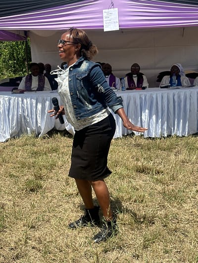 A woman in a denim jacket, white shirt, and black skirt is holding a microphone, speaking into it. She is wearing glasses, and her hair is pulled back in a ponytail. She is standing in a grassy area in front of a white tent. The tent has a purple striped awning and a white sign hanging from it. There is a table with a white tablecloth behind her, where multiple people are seated. The location is in Pridelands, Kenya, based on the coordinates -1.302, 37.02. The environment is dry and sunny.