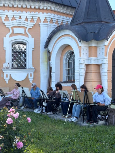 Six people are seated on chairs in front of an ornate, peach-colored building with white decorative elements. The building appears to be a historical church or monastery. All six individuals are engaged in painting or drawing, with easels and canvases set up before them.
From left to right:
The first person is a woman wearing a pink hoodie and a dark skirt. She is looking at her canvas and appears to be actively painting.
The second person is a man wearing a blue denim shirt, a baseball cap, and blue jeans. He is looking down at his canvas.
The third person is a woman with curly dark hair, wearing a plaid shirt, and looking at her canvas.
The fourth person is a young woman with long dark hair, wearing a plaid shirt, and focused on her artwork.
The fifth person is a young woman with dark hair, wearing a light-colored hoodie and jeans, looking intently at her phone, which is resting on her easel.
The sixth person, a woman wearing a red baseball cap and a light-colored hoodie, is also looking at her phone.
In the foreground, to the left, there are several large pink peonies and green foliage. The ground in front of the building is paved with stone, and beyond that is a grassy area. The overall impression is of an outdoor art class or plein air painting session in front of a significant architectural structure in the city of Suzdal, Russia.