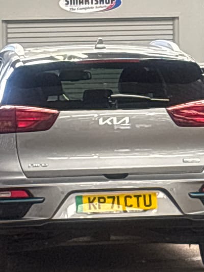 The rear of a silver Kia Niro electric vehicle is visible, parked outdoors. The car has a black rear spoiler and a shark fin antenna. The Kia logo is centered on the tailgate. A yellow license plate with green lettering reads "KP71 CTU". The car is positioned in front of a grey corrugated metal garage door. Above the garage door, a white oval sign with blue text that appears to say "SMARTSHOP The Complete Solution" is visible. The scene is likely located in an industrial or commercial area within the city of Derby, United Kingdom, given the presence of the "SMARTSHOP" sign which is a known retailer in Derby. There are no people visible in the image.