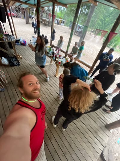 In Luzhki, Russia, a group of people are gathered on a wooden deck beneath a thatched roof structure. The scene appears to be outdoors, with trees and a sandy area visible in the background. A man in the foreground is taking a selfie, wearing a red tank top and a green glow necklace. Other individuals are seen interacting, some sitting at tables with drinks and food, while others are standing and talking. The atmosphere suggests a casual gathering or party.