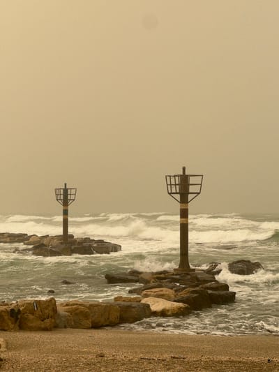 This image depicts a turbulent coastal scene in Jisr az-Zarqa, Israel, under an overcast, yellowish-brown sky indicative of atmospheric haze or a dust event. Two prominent pole-like navigational markers are positioned on separate rocky breakwaters extending into the sea. The marker on the left exhibits a dark vertical shaft with green and dark horizontal stripes near its base, topped by a dark, open-frame structure. The marker on the right, also dark, has red and dark horizontal stripes near its base and a similar dark, open-frame top.
The sea is rough, with numerous white-capped waves crashing against the dark, irregular rocks forming the breakwaters. The water displays a greenish-blue hue amidst the white foam. In the immediate foreground, a narrow strip of coarse, light brown and grey pebbly beach is visible. No individuals or other active subjects are present in the scene.