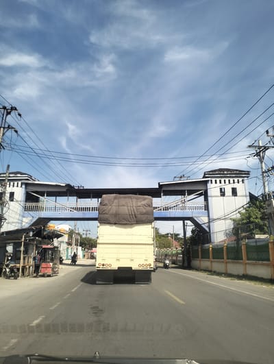 A wide-angle shot depicts a truck on a road, seen from the perspective of a vehicle behind it. The truck, a pale yellow box truck with a brown tarp covering its cargo, is centrally positioned and moving away from the camera. The road is asphalt, marked with a yellow dashed line down the center and white dashed lines on the edges.

Above the road, a pedestrian bridge spans from one side to the other, supported by two distinct structures. These structures are white with dark roofs and blue railings on the bridge itself. Numerous electrical wires crisscross the sky, originating from utility poles visible on both sides of the road.

To the left of the road, several small market stalls are set up. A few people are visible, including someone standing near a stall and others on motorcycles. To the right, a green fence runs along the edge of a property, with some trees visible behind it.

The sky is a vibrant blue, dotted with white, wispy clouds, suggesting it is daytime, likely mid-morning or afternoon. The overall scene is of a busy, somewhat cluttered street in what appears to be a town or city in Indonesia. The presence of market stalls, utility poles, and the architectural style of the bridge supports suggest a tropical or sub-tropical location. No discernable text is visible in the image.