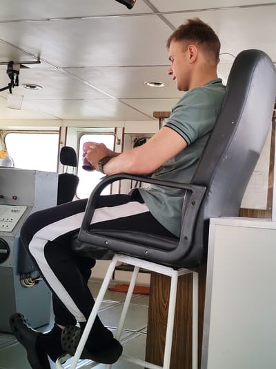 A man is seated in a high-backed, black, swivel chair with armrests inside what appears to be a control room on a vessel, possibly a ship or boat. The man, who has short blond hair, is wearing a green polo shirt with black stripes at the collar and sleeves, black track pants with a white stripe down the side, and black Crocs. He is holding a purple mug in his hands.

The control room has a gray console with various switches and gauges to the left of the man. Through a window behind him, there is bright daylight. The room has a white, panelled ceiling with recessed lighting. Based on the description, the location is in the city of Приволжский, Russia.