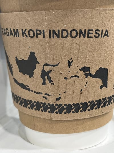 The image shows a close-up of a brown paper coffee cup sleeve with a black stencil of the Indonesian archipelago printed on it. The text "RAGAM KOPI INDONESIA" is printed in black capital letters above the map. The coffee cup itself is white and is partially visible at the bottom of the frame. The background is out of focus and appears to be a light-colored surface. There are no people or animals visible in the image. The scene is indoors and appears to be well-lit. There is no indication of the specific location within Singasari, Indonesia, other than the text on the cup sleeve.