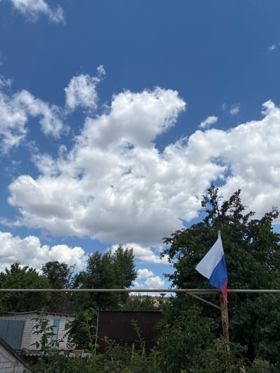 The image depicts a view of a clear blue sky with scattered white clouds. In the foreground, a Russian flag is displayed, attached to a rustic pole. The flag is partially unfurled, showing its white, blue, and red colors. Behind the flag, lush green trees dominate the scene, with their leaves forming a dense canopy. Below the trees, various structures are visible, including a white brick building with a blue door, suggesting a residential or outbuilding. A metal pipe or railing runs horizontally across the mid-ground. The overall setting appears to be a rural or suburban area, possibly a backyard or a communal space within Zhirnov, Russia. The lighting indicates it is daytime, with the sun casting natural light on the scene.