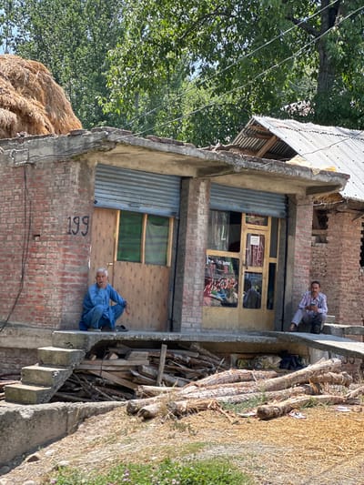 In Vessu, India, a man in blue traditional clothing sits squatting on a concrete ledge in front of a brick building. The building has a corrugated metal shutter pulled down over a window and a wooden door with glass panels. Next to him, a stack of hay is piled on the roof. To the right of the building, another man in a checkered shirt and light trousers sits on the steps of a neighboring structure, looking at his phone. The ground in front of the buildings is covered in dry straw and scattered logs, with some greenery visible in the foreground.