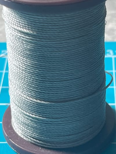 A close-up, eye-level, vertical shot shows a spool of teal-colored thread. The spool has dark brown, circular caps on the top and bottom. The thread is wound tightly around the spool, creating horizontal ridges. The background is out of focus but appears to be a blue grid-like surface, possibly a cutting mat. A single strand of thread extends outward from the spool on the right side. The scene is lit from above and slightly to the left, casting subtle shadows. There are no people, animals, or recognizable landmarks in the image. The location in Novy Urengoy, Russia, is not discernible from the visual information presented.