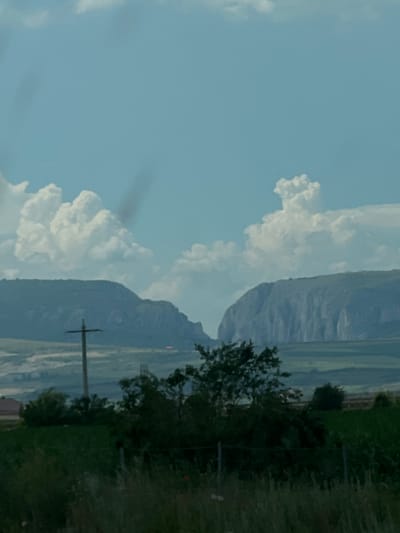 The image shows a landscape with large rock formations in the background, possibly mountains or mesas. The sky is blue with scattered white clouds. In the foreground, there are green fields and trees. A telephone pole with wires is visible to the left of the center. The overall scene suggests a rural or natural environment. The specific location in the city of Mihai Viteazu, Romania is not discernible from the image itself, as there are no landmarks or identifying features that place it there.