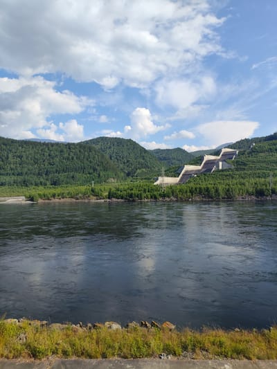 The image shows a wide, dark river in the foreground. The river is calm with ripples and reflections of the sky. Along the riverbank, there is a strip of green grass and rocks. In the middle ground, there is a dense forest covering rolling hills. A large, stepped concrete structure, identified as the Sayano-Shushenskaya Hydroelectric Power Station, is built into the hillside on the right side of the frame, surrounded by trees. Power lines and pylons are visible near the structure. In the background, more green, forested mountains rise up, with some parts fading into the distance. The sky is blue with scattered white, fluffy clouds, suggesting a clear day. The scene is set in the town of Cheremushki, Russia, a location known for the Sayano-Shushenskaya Dam. There are no people or animals visible in the image.