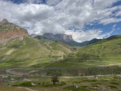The image depicts a panoramic view of a mountain valley in El'tiubiu, Russia. The landscape is characterized by rolling green hills, rugged mountain peaks, and a winding asphalt road that snakes through the valley. The sky is partially overcast with dramatic clouds interspersed with patches of blue. In the foreground, there are grassy slopes dotted with rocks and a few scattered trees. The middle ground features the road, which appears to lead to small clusters of buildings and structures, including what look like agricultural buildings or farmsteads. The background is dominated by imposing mountain ranges, some of which are shrouded in mist or clouds. The overall scene conveys a sense of remote natural beauty and rural development. There are no visible people or animals in the image. The environment is clearly mountainous and rural, likely at a moderate elevation given the vegetation.