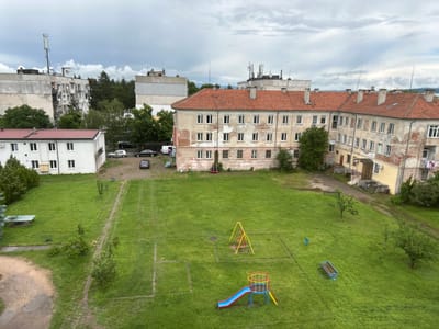 This overhead view depicts a residential area in Gara Elin Pelin, Bulgaria, on an overcast daytime. The scene is dominated by several multi-story buildings and a large, central grassy courtyard.
In the foreground and center, a well-maintained green lawn covers most of the space. Faint, worn-out white lines suggest a former sports field or play area. On the grass, there's a small children's playground featuring a colorful blue and red slide attached to a circular platform, and a yellow swing set. A blue park bench and a blue trash can are also visible on the lawn. Several small trees and bushes are scattered throughout the grassy area. Dirt paths and tire tracks cut across the lawn, leading towards buildings and parking spots.
Surrounding the courtyard are various residential buildings. To the left, a modern-looking white two-story building with a reddish-brown roof and multiple windows is visible. Behind it, partially obscured by trees, are taller, older apartment blocks with exposed concrete and numerous satellite dishes on their balconies and roofs. Prominently in the midground and right, an older, L-shaped building with a distinctive red tiled roof and a worn, peeling facade stands. This building features multiple windows and several chimneys. Its walls show signs of age and neglect, with large patches of plaster missing, revealing brickwork. Laundry is seen drying on a line outside one of its ground-floor windows.
The sky is cloudy and grey, suggesting an overcast day without direct sunlight. Several cars and a white van are parked on dirt and asphalt patches near the buildings. The roofs of the taller buildings feature various antennas and cellular towers. No people are visible, lending a quiet, still atmosphere to the scene. There is no discernible text visible on any of the buildings, vehicles, or objects.