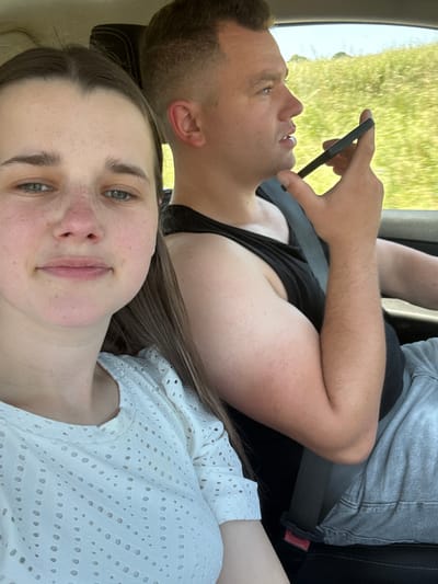 A woman, who is facing the camera, and a man are inside a vehicle. The woman has light skin, is wearing a white shirt, and has brown hair. The man, on the right side of the image, is wearing a black tank top and gray pants, and is holding a black smartphone to his ear. The vehicle appears to be moving through a rural area with green fields and trees visible through the windows. The location is in Niederwetz, Germany.