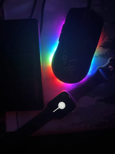 The image displays a close-up, top-down view of electronic devices on a surface in a dimly lit indoor environment, illuminated by multicolored LED lighting. The specific location in Ribnovo, Bulgaria, cannot be determined from the visual information provided.
From left to right and top to bottom:
- **Left side:** A dark, rectangular electronic device, possibly a laptop or tablet, is oriented vertically. Its screen is dark.
- **Top center:** A dark computer mouse with a visible cord extends upwards. The mouse features a repeating hexagonal or honeycomb pattern of cutouts on its surface.
- **Bottom right:** A dark-colored smartwatch, consistent with an Apple Watch, is positioned diagonally. Its rectangular screen is illuminated, displaying a white magnifying glass icon with a circular center on a black background, indicating a search function or similar status. The watch band is also dark-colored and appears to have a textured pattern.
- **Environment:** The devices rest on a surface, likely a desk or mouse pad, which exhibits a subtle, dark pattern resembling foliage or trees on the right side. Behind the devices, a strip of multicolored LED lighting casts a radiant glow, transitioning through red, orange, green, blue, and purple hues against a dark background, possibly a wall or panel.
No individuals are present, and no actions or interactions are depicted. The scene suggests a personal computing setup in low ambient light conditions.