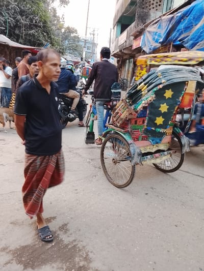 The image depicts a bustling street scene in Tarabo, Bangladesh. In the left foreground, a man with a thin build, short dark hair, and a light complexion stands facing slightly right, looking forward. He wears a black short-sleeved polo shirt with white trim on the sleeves and a red-patterned lungi, secured at the waist, reaching his ankles. Dark open-toed sandals are on his feet, and his hands are clasped behind his back. A brown dog is partially visible behind his left leg.
To the right of the foreground man, a brightly colored, three-wheeled bicycle rickshaw is parked or moving slowly. Its passenger carriage is highly decorated with intricate paintwork and patterns, including three prominent yellow stars on a dark green section of its folding canopy. The rickshaw's frame and wheels show signs of use. A man is partially visible behind the rickshaw, appearing to be operating it; he wears a dark long-sleeved jacket, light blue jeans, and dark sandals, his back mostly to the viewer.
In the midground, multiple individuals and vehicles navigate the street. To the left, a group of men stands near what appears to be a market stall, one wearing a white and yellow checkered lungi. Further back, a man on a black motorcycle, wearing a helmet, drives away from the camera. Another person on a bicycle is visible behind the rickshaw operator.
The background features multi-story buildings, some with blue tarpaulins draped from their facades, suggesting shops or temporary shelters. Dense green foliage from trees is visible on the far left. Utility poles and overhead lines are present throughout the background. The street surface is paved, appearing somewhat worn, with scattered dark stains. The overall lighting suggests daytime.