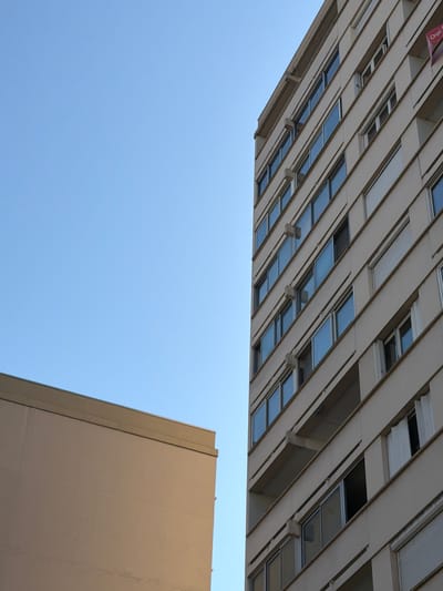 The image shows an urban scene with two buildings against a clear blue sky. The building on the right is a tall, multi-story apartment building with a light-colored facade. It features a repeating pattern of windows with dark blue or black frames, and some of these windows have visible blinds or curtains. The building on the left is shorter and has a plain, tan-colored facade. The perspective suggests the viewer is looking up at the buildings from a street or lower vantage point. A small red and white sign is visible at the top right corner of the apartment building, partially obscured by its angle. The scene is devoid of people or any immediate signs of human activity beyond the architectural structures themselves. The overall impression is one of a typical residential area in a city.