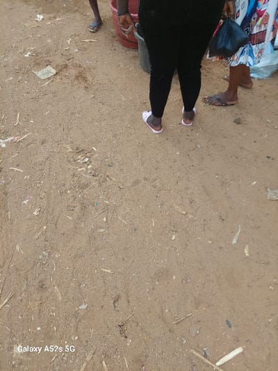 The image portrays an unpaved outdoor area in Awkuzu, Nigeria; the precise location within the city is not discernible. The ground is composed of light brown, sandy soil interspersed with debris including small pieces of plastic, organic matter, and a crumpled piece of paper currency in the upper left quadrant. Three individuals are visible from the knees downward, all exhibiting dark brown skin tones. The person on the far left wears blue flip-flops. The central individual wears black pants and white sandals, holding a dark object in their right hand near a red plastic basin and a dark grey bucket. The person on the right wears brown flip-flops and a patterned garment, carrying a black plastic bag in their left hand. All individuals are depicted in a standing posture.