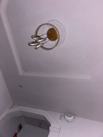 A low-angle shot reveals a white ceiling with decorative molding. A modern chandelier with gold accents and silver, swirling elements hangs from a circular medallion. A faint crack runs across the ceiling's surface. In the upper right, a white, decorative plate is mounted near a doorway with ornate molding and a glimpse inside reveals a dark interior space.