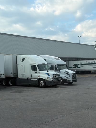 Two white semi-trucks are parked in front of a large warehouse building. The truck on the left has a white trailer and a white cab with dark gray trim. The truck on the right has a white trailer with "SCT Southern Chain" and "Paperboard Packaging" printed on it in green and black lettering, and a white cab with dark gray trim. The warehouse has a corrugated metal exterior and several loading docks marked with numbers 45, 46, 47, and 48. A light pole with two lights and an antenna is visible on top of the warehouse. The sky is overcast with light clouds. The ground is asphalt, with some cracks and water spots visible. The scene is forensic and matter-of-fact.