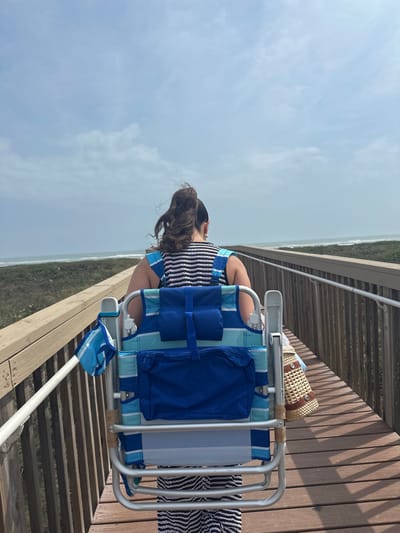An individual, seen from behind, is walking forward on a wooden boardwalk in South Padre Island, United States of America. The person has dark hair styled in a high ponytail and is wearing a black and white striped sleeveless top and coordinating lower garment. A foldable beach chair, featuring blue, light blue, and white stripes with dark blue pockets, is strapped to the person's back, serving as a backpack. A small blue and white striped pouch hangs from the left side of the chair, while a light brown woven straw bag with dark circular patterns hangs from the right side. The boardwalk is constructed from brown wooden planks and features wooden railings on both sides, with an additional white handrail on the left. Low coastal vegetation flanks the boardwalk. In the background, beyond the vegetation, the ocean with visible waves is present under a partly cloudy sky.
