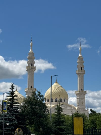 The image shows a white mosque with a golden dome and two minarets against a blue sky with white clouds. The mosque is partially obscured by trees in the foreground. A yellow sign with black lettering and a pedestrian crossing sign are visible on the left side of the image. The scene is outdoors on a sunny day. This is the White Mosque in Bolgar, Tatarstan, Russia, which is located on the territory of the ancient city of Bolgar.