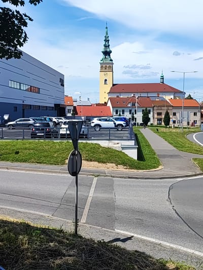 This media file captures a daytime scene in Nový Jičín, Czechia, under a bright, partially cloudy sky, suggesting fair weather. The foreground is dominated by a paved road with lane markings and a grassy verge with dry grass. A black metal pole with two circular signs is positioned near the intersection of two roads.
Further back, a parking lot with several cars is visible, separated from the foreground by a black metal fence and a strip of green grass. The cars appear to be parked in an organized manner. Beyond the parking lot, a modern, light-colored building with horizontal window elements stands to the left.
The background features a cluster of buildings with red-tiled roofs, characteristic of European architecture. Prominently, a tall, yellow church tower with a green spire rises above the other structures. A clock face is visible on the tower. Another smaller green-spired structure is also present. A person can be seen walking on a paved path that leads between the parking lot and the buildings. A streetlamp is also visible. The overall impression is of an urban or town setting with a mix of modern and historical architecture.